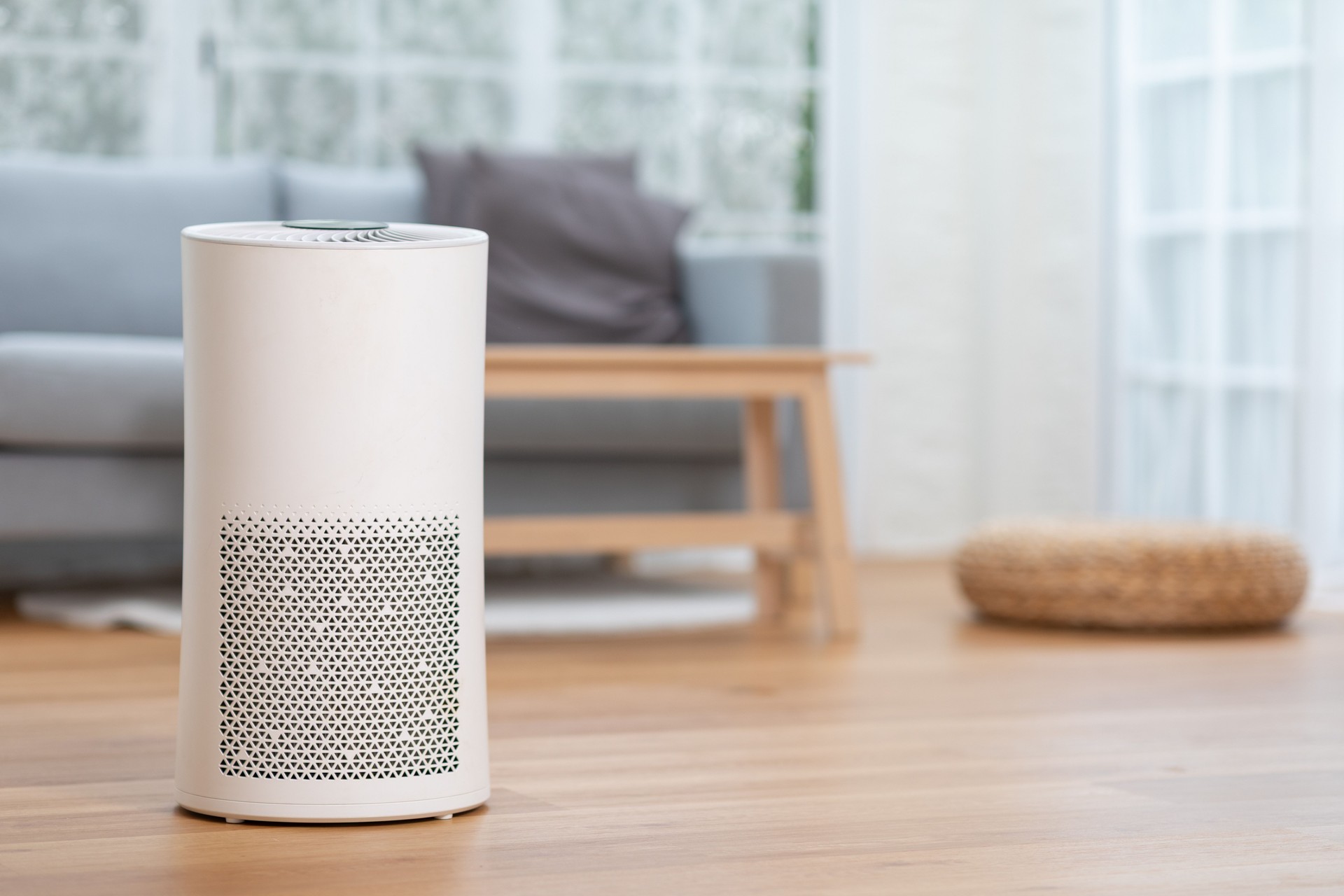 modern white air purifier placed on wooden floor in bright, cozy living room with sofa, cushions, and natural light streaming through large windows